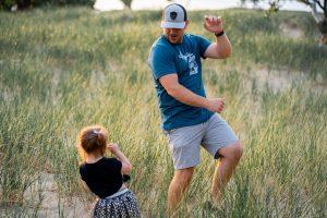 man in blue shirt and grey shorts with daughter dancing in a field with long grass